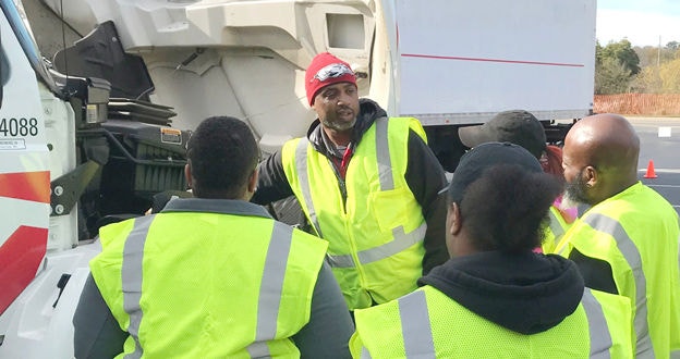 Manager training small group of drivers while pointing to the engine area or a large truck. All people in hi-vis safety vests