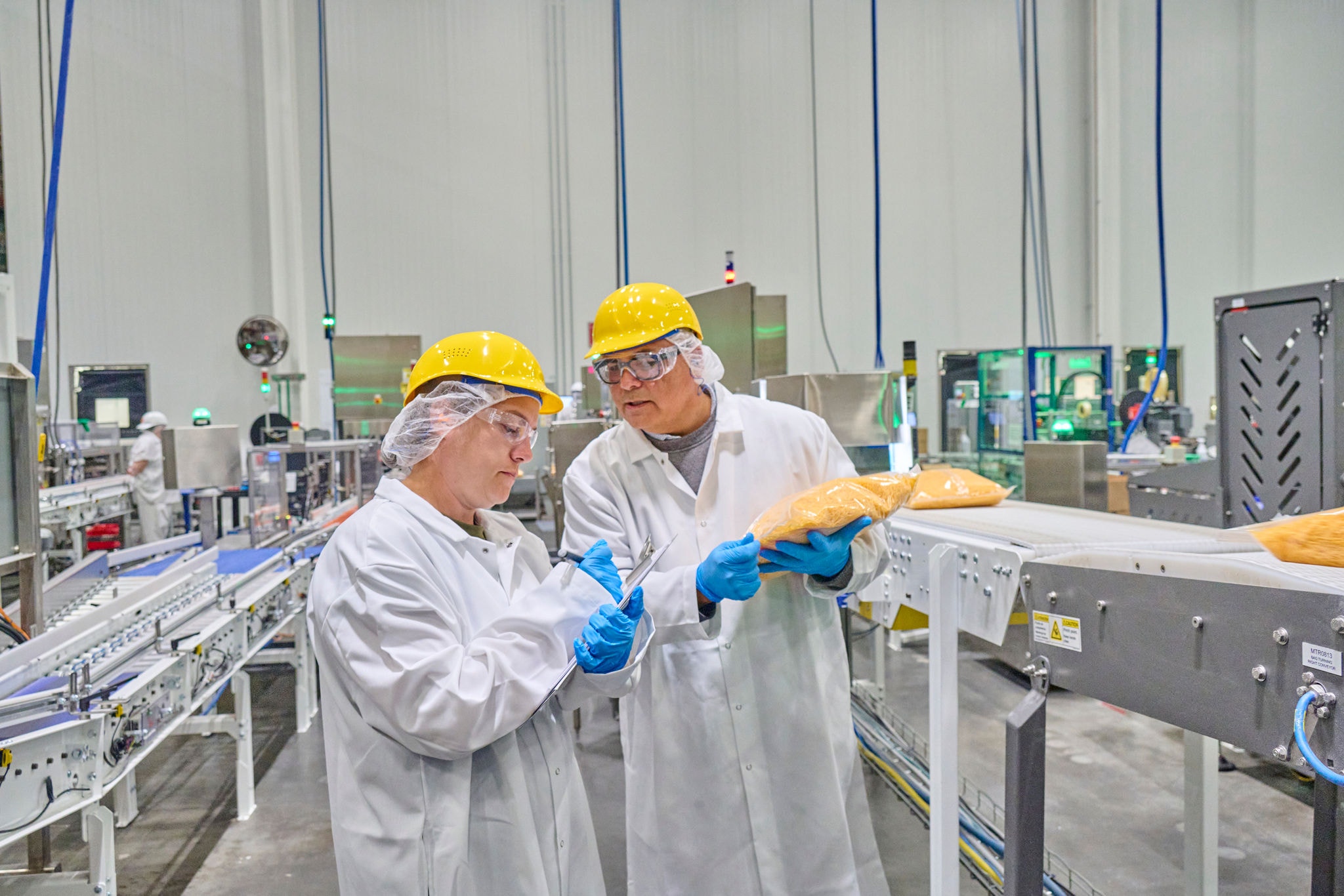 Two people in cheese processing facility looking at a clipboard while examining a bag of shredded cheese